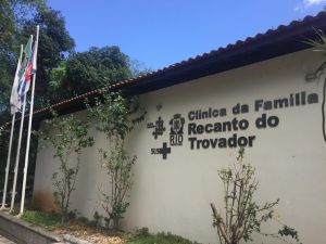 Imagem exibindo a fachada da Clínica da Família Recanto do Trovador. É mostrado um prédio de um unico andar, com telhado de madeira e uma parede bege exibindo o nome da clínica em primeiro plano Autoria: Clínica da Família - Recanto do Trovador