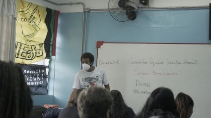 Em destaque, um homem negro de máscara que está de frente para a turma. Atrás dele há um quadro branco, com algumas anotações e à sua esquerda duas bandeiras cobrindo a janela.