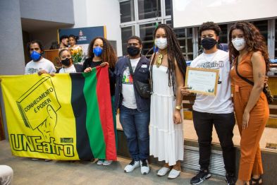 Sete jovens lado-a-lado dentro do auditório da Alesp. À esquerda, três deles seguram uma bandeira da UNEafro com a cor amarelo de fundo e faixas de cor preta, vermelha e verde. Ainda na bandeira, uma mão segura um livro que leva os dizeres "Conhecimento é poder".À direita, um dos jovens segura uma placa de honraria.